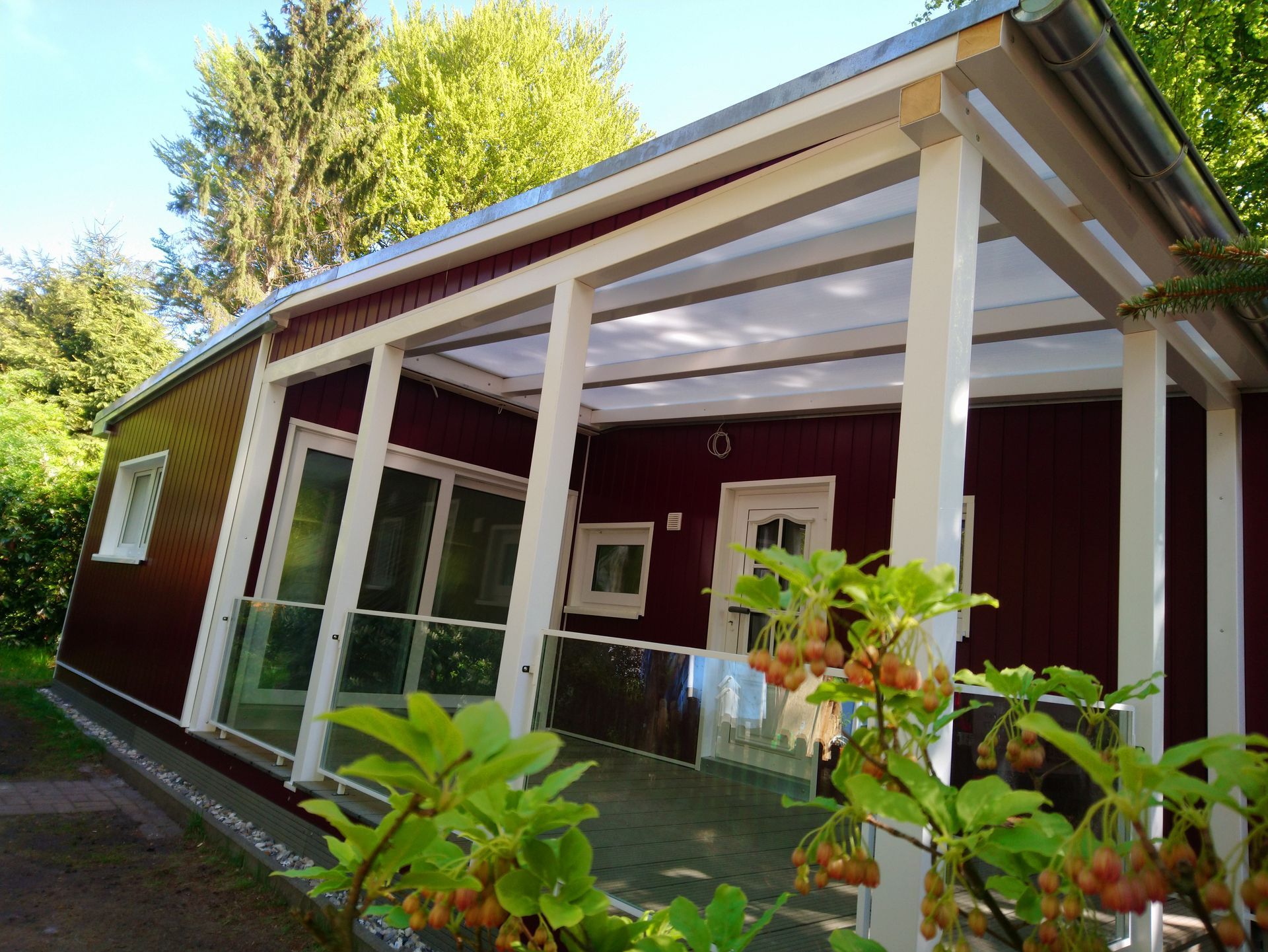 Rote Hütte mit weißer Veranda und Glasgeländer. Grüne Bäume umgeben die Hütte. Sonniger Tag.