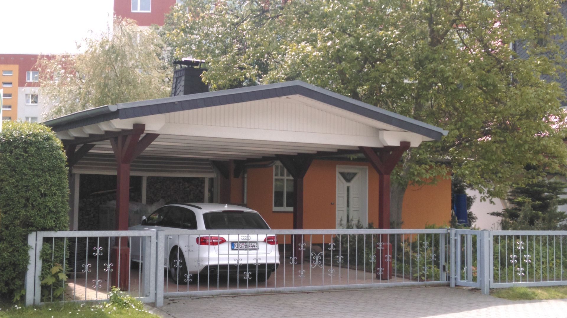 Ein weißes Auto parkt unter einem Carport, der an ein orangefarbenes Haus angebaut ist, hinter einem Metalltor.