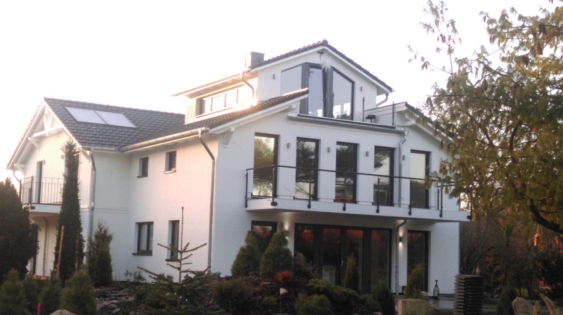 Weißes, zweistöckiges Haus mit großen Fenstern, Balkonen und einem schrägen Dach. Sträucher und Bäume umgeben das Haus.