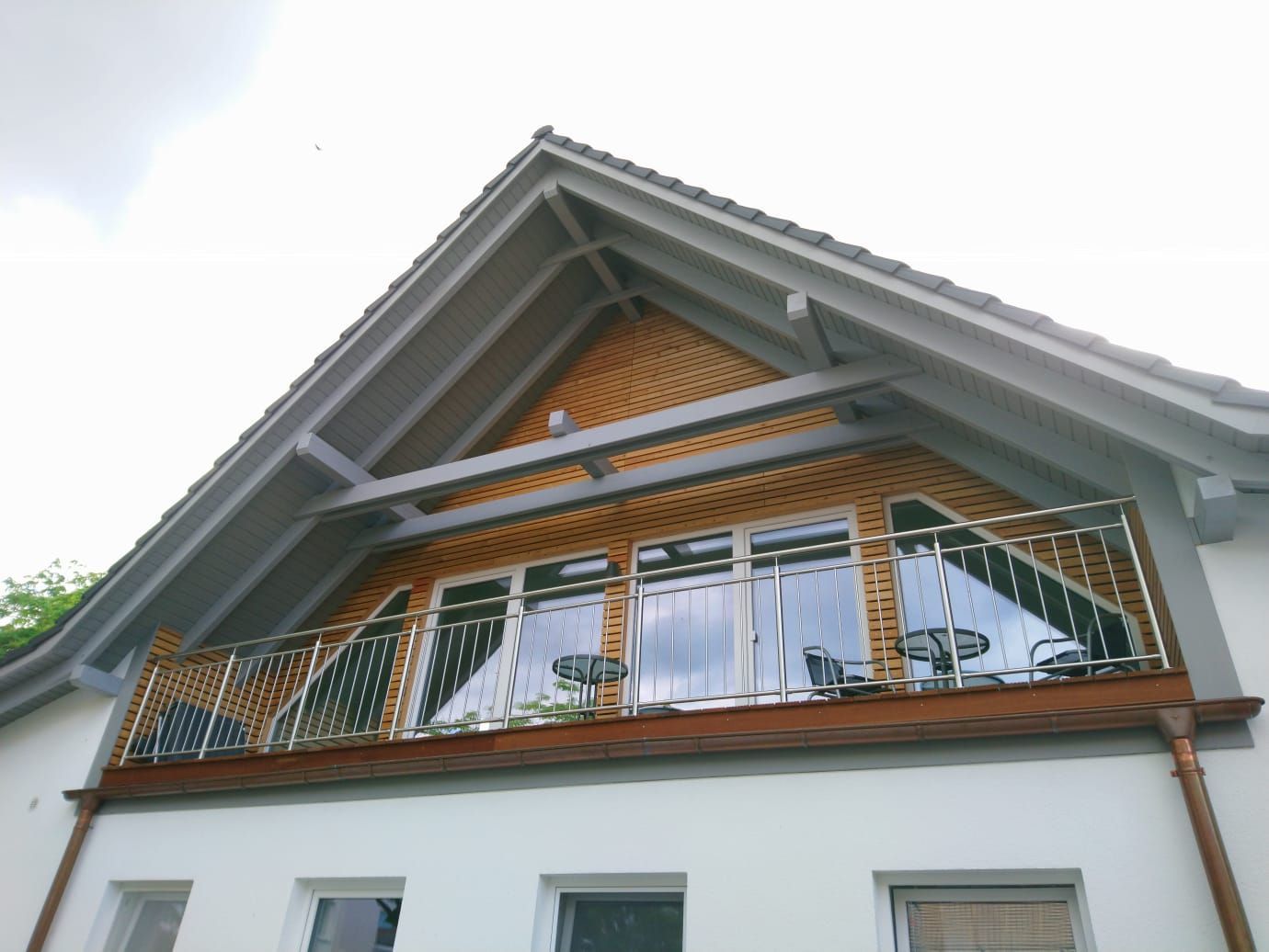 Balkon mit Glastüren, Metallgeländer, Holzelementen und schrägem Dach an einem weißen Gebäude.
