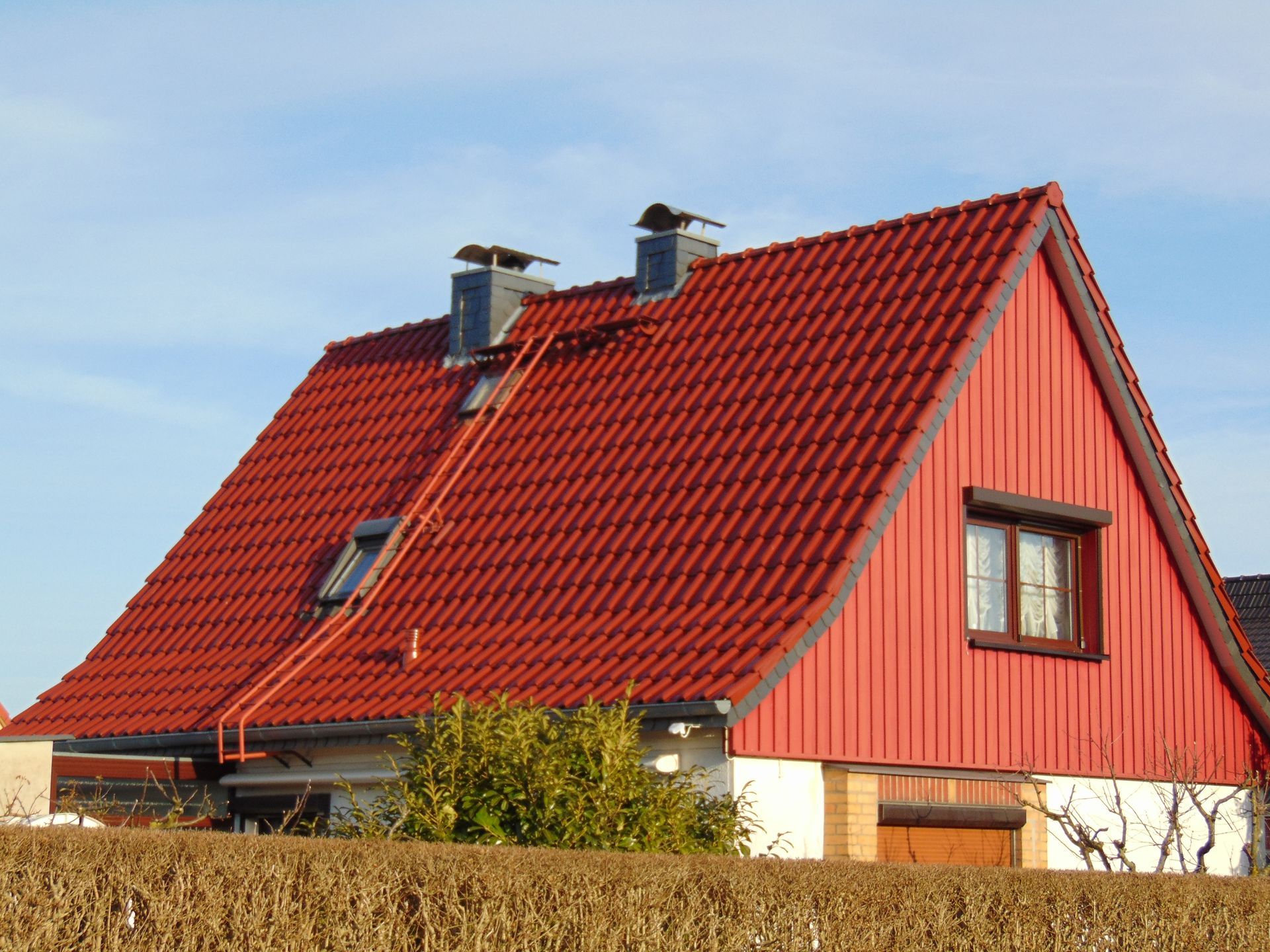 Haus mit rotem Dach und roter Fassade, zwei Schornsteinen und einer Leiter am Dach.