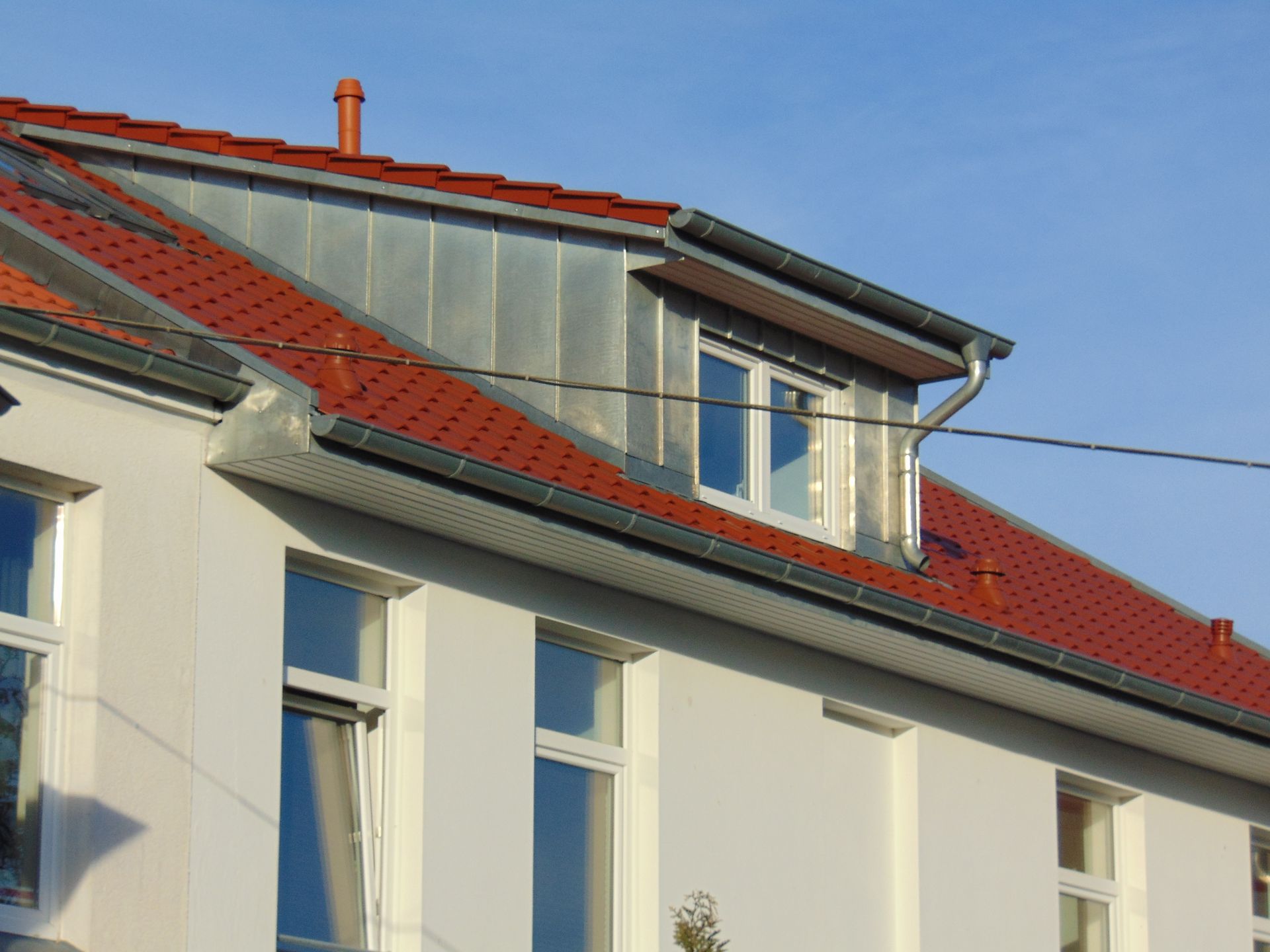 Weißes Gebäude mit rotem Ziegeldach und metallener Dachgaube vor blauem Himmel.