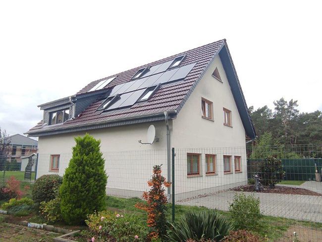 Zweistöckiges Haus mit Solaranlage auf dem Dach und Satellitenschüssel. Grüner Garten und bewölkter Himmel.