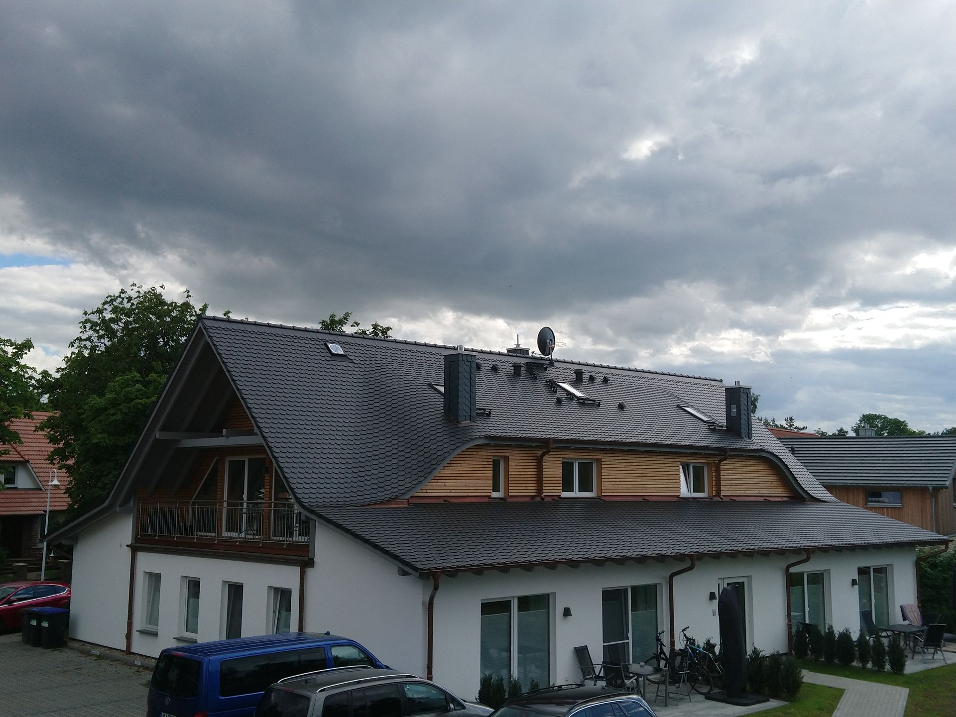 Weißes Haus mit dunklem Dach und Holzelementen unter bewölktem Himmel. Autos parken davor.