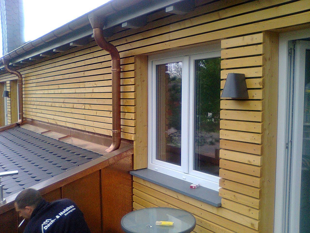 Außenansicht eines Gebäudes mit Holzverkleidung, weißgerahmten Fenstern, Kupferrinnen und einer Person, die auf der Terrasse arbeitet.