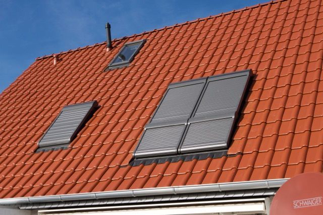 Rotes Ziegeldach mit mehreren Oberlichtern und grauen Fensterläden vor blauem Himmel.