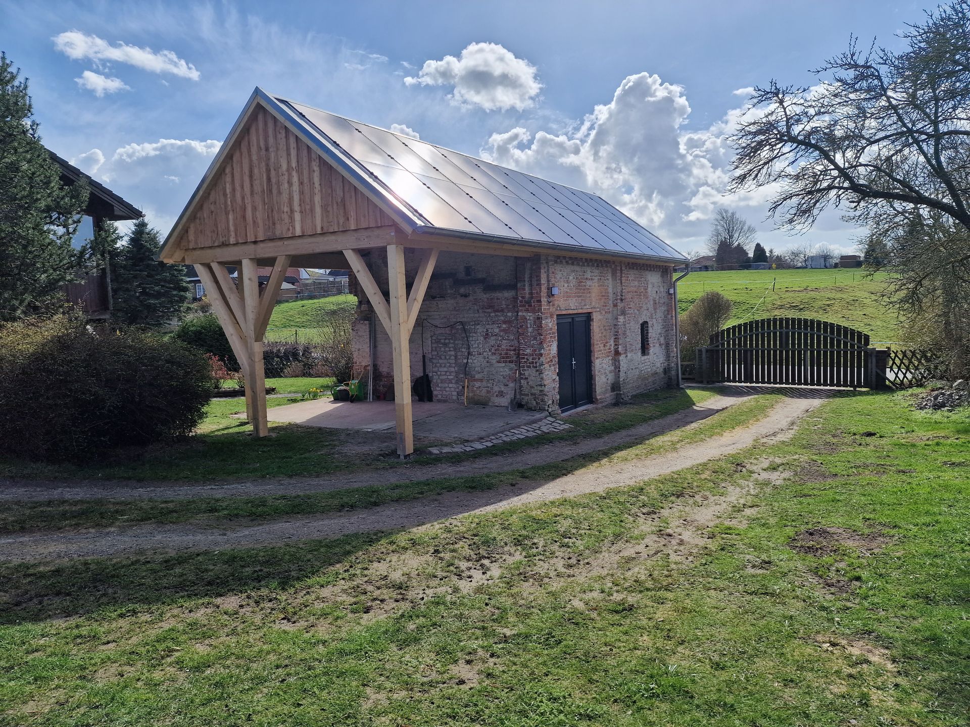 Backsteingebäude mit Holzveranda, Metalldach und Auffahrt auf einer grasbewachsenen Fläche mit Tor und Bäumen.