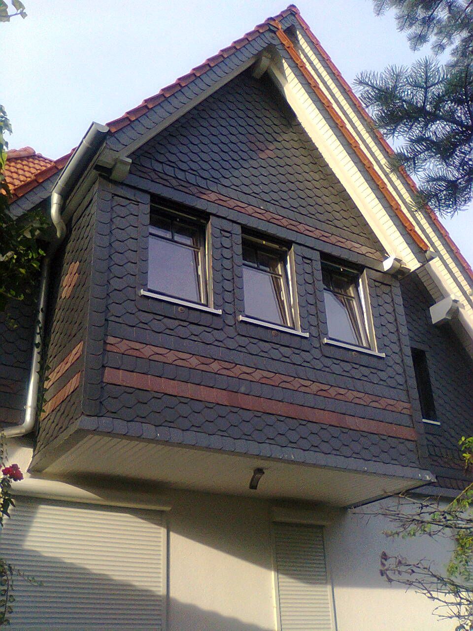 Erkerfenster mit dunkler Schieferverkleidung und drei Fenstern; an einem hellen Gebäude mit rotem Ziegeldach.