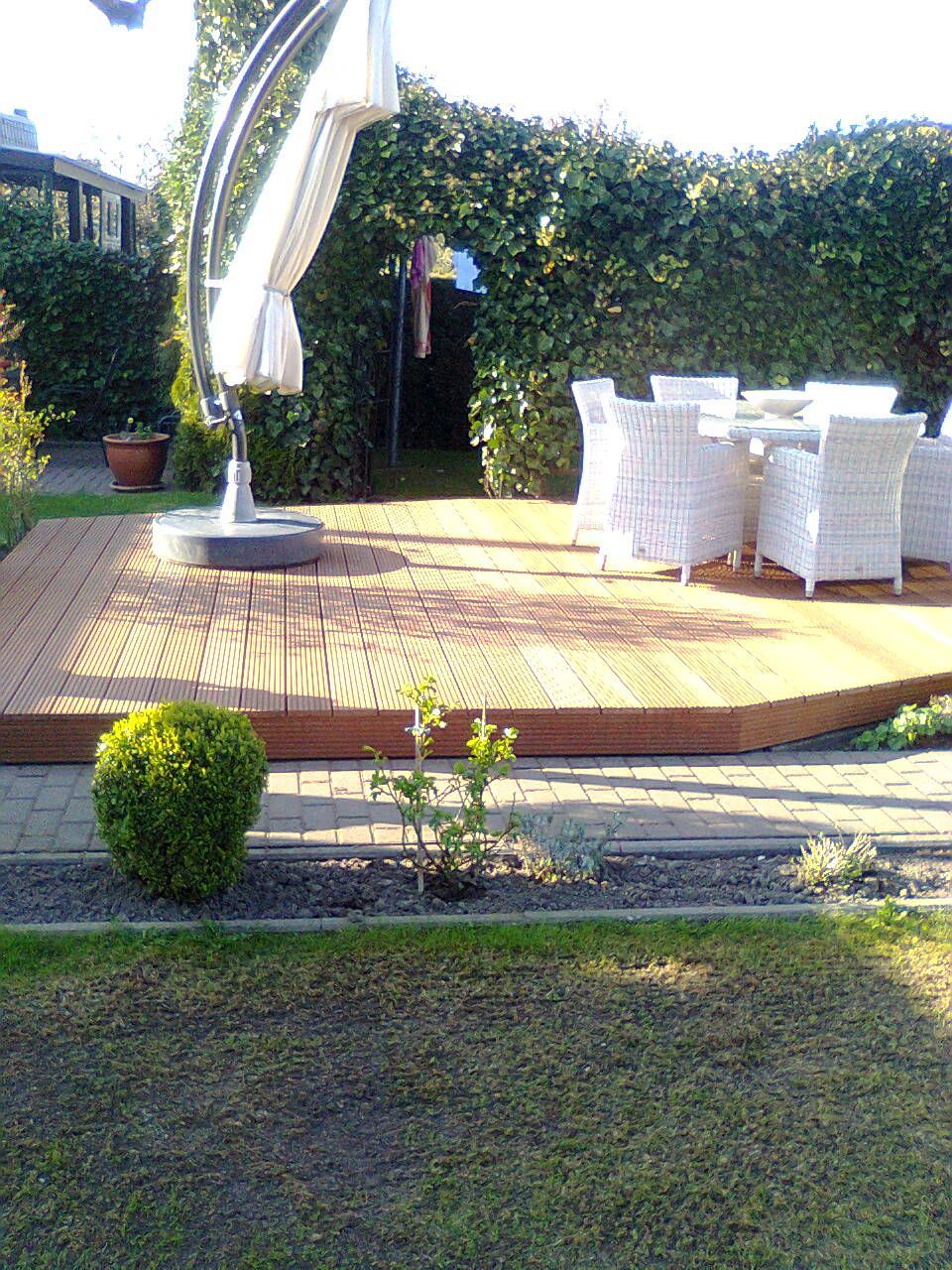 Holzterrasse mit Essgruppe unter einem Sonnenschirm, im Hintergrund üppiges Grün.