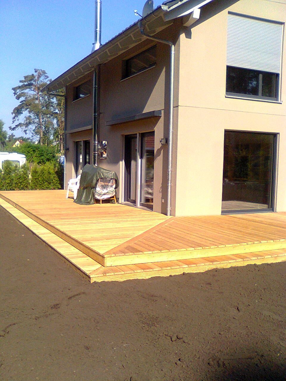 Holzterrasse vor einem zweistöckigen Gebäude mit großen Fenstern und einem Schornstein. Braune Fassade, sonniger Tag.
