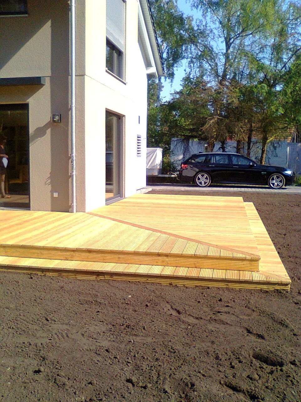 Holzterrasse mit Treppe neben einem beigen Haus, im Hintergrund ein geparktes Auto an einem sonnigen Tag.