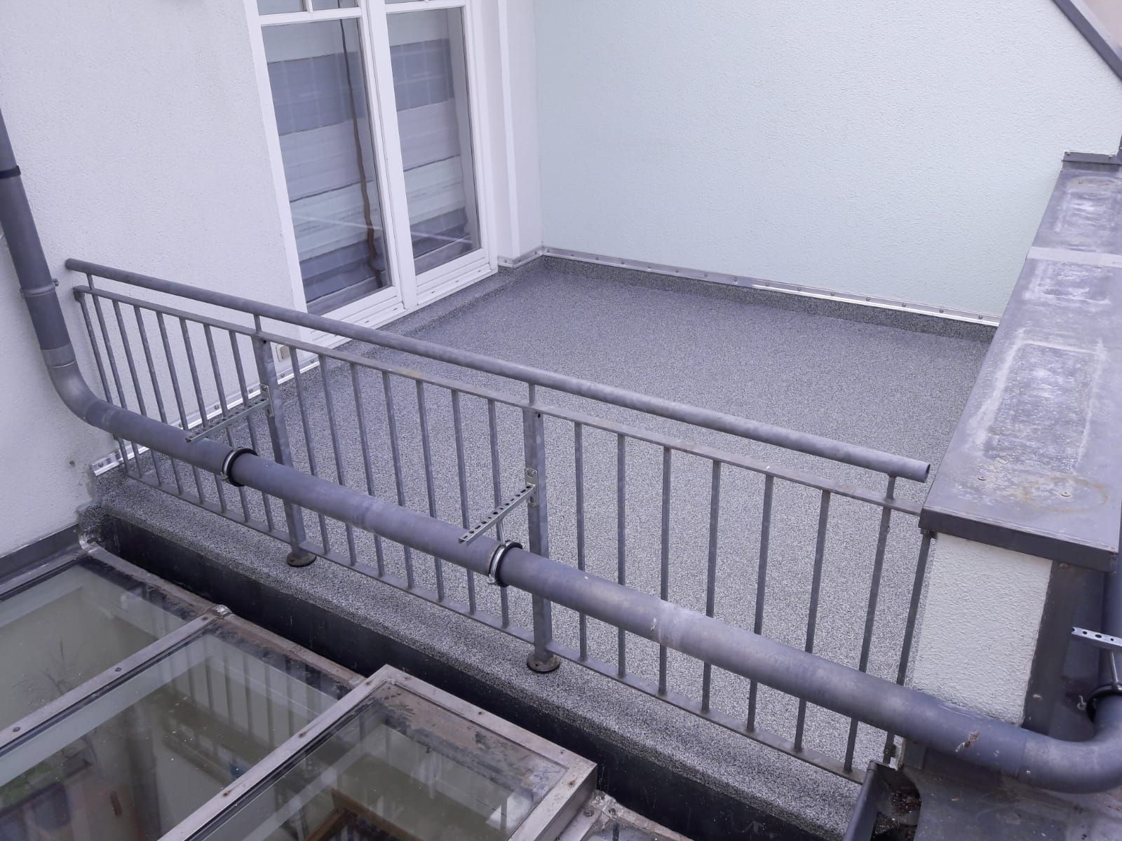 Balkon mit grauem Bodenbelag, Metallgeländer und weißer Wand, neben Glasdachfenstern und einem Fallrohr.