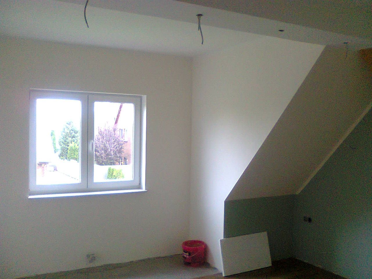 Innenansicht eines im Bau befindlichen Raumes mit Fenster, weißen Wänden und schräger Decke.