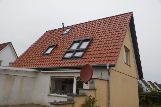 Haus mit orangefarbenem Ziegeldach, Oberlichtern und Satellitenschüssel vor bewölktem Himmel.