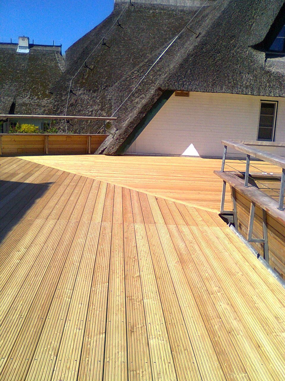 Holzterrasse mit Strohdach im Hintergrund. Sonnenlicht wirft Schatten auf die Oberfläche der Terrasse.