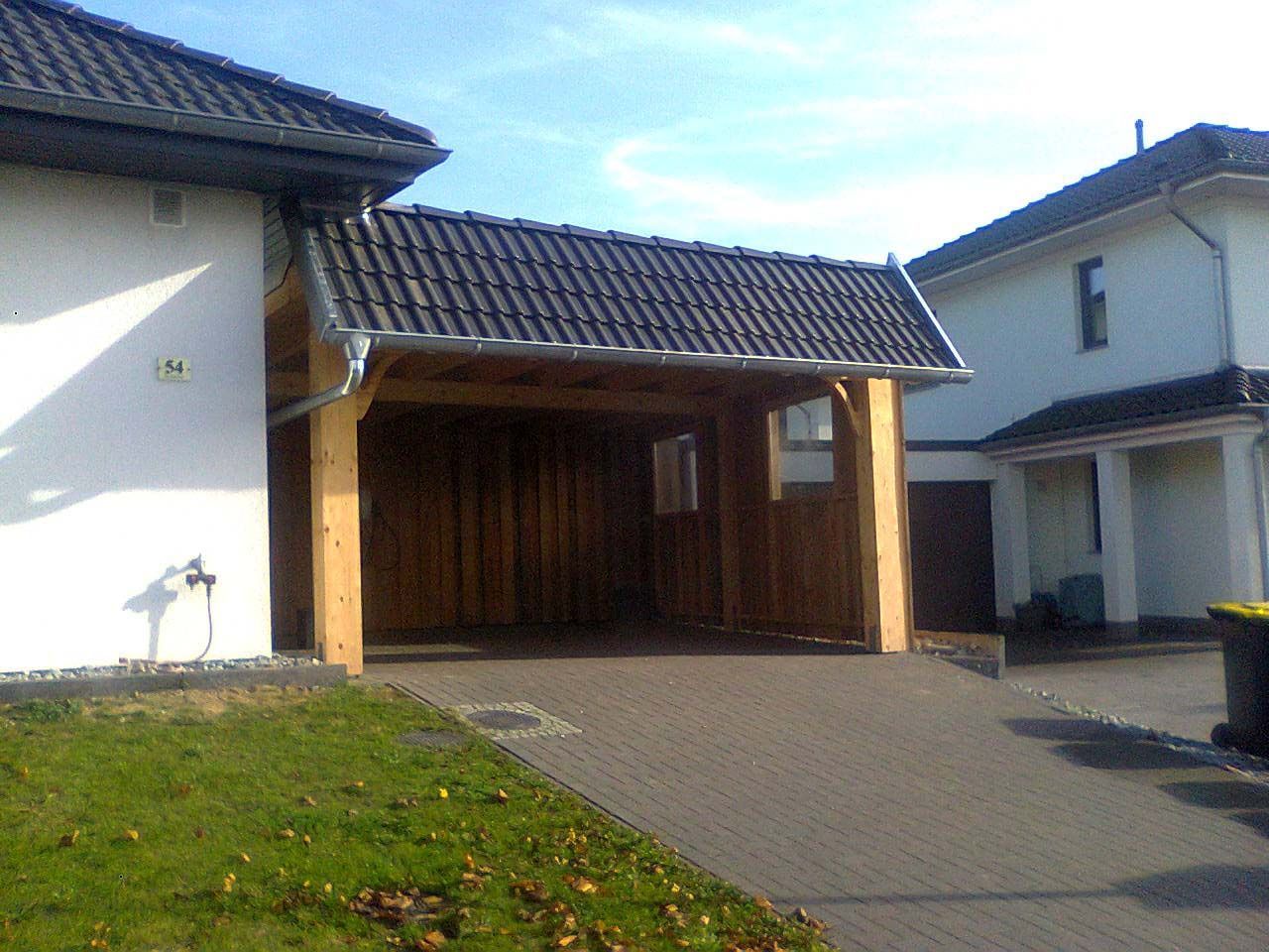 Ein hölzerner Carport, der an ein weißes Haus mit gepflasterter Auffahrt und grünem Rasen an einem sonnigen Tag angrenzt.