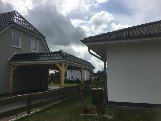 Carport an grauem Haus mit dunklem Dach; Holzstützen. Bewölkter Himmel.