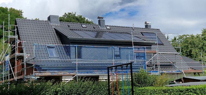 Haus mit dunkelgrauem Dach, im Bau. Das Gebäude ist von einem Gerüst umgeben, Solarpaneele sind installiert.