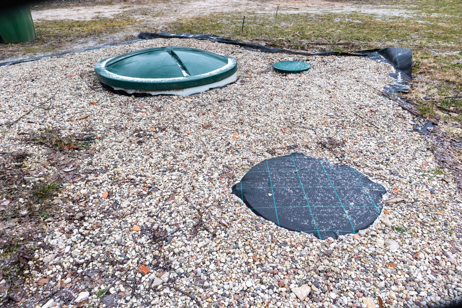 Trois couvercles de fosses septiques posés sur du gravier, à l'extérieur. Un grand, deux petits.