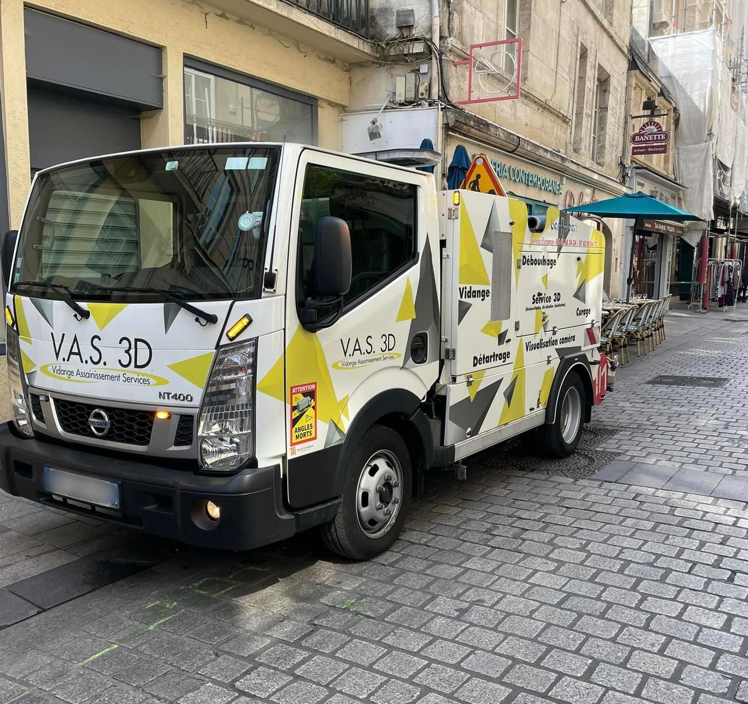 Un camion Nissan blanc portant une inscription est stationné dans une rue pavée, près d'un bâtiment.