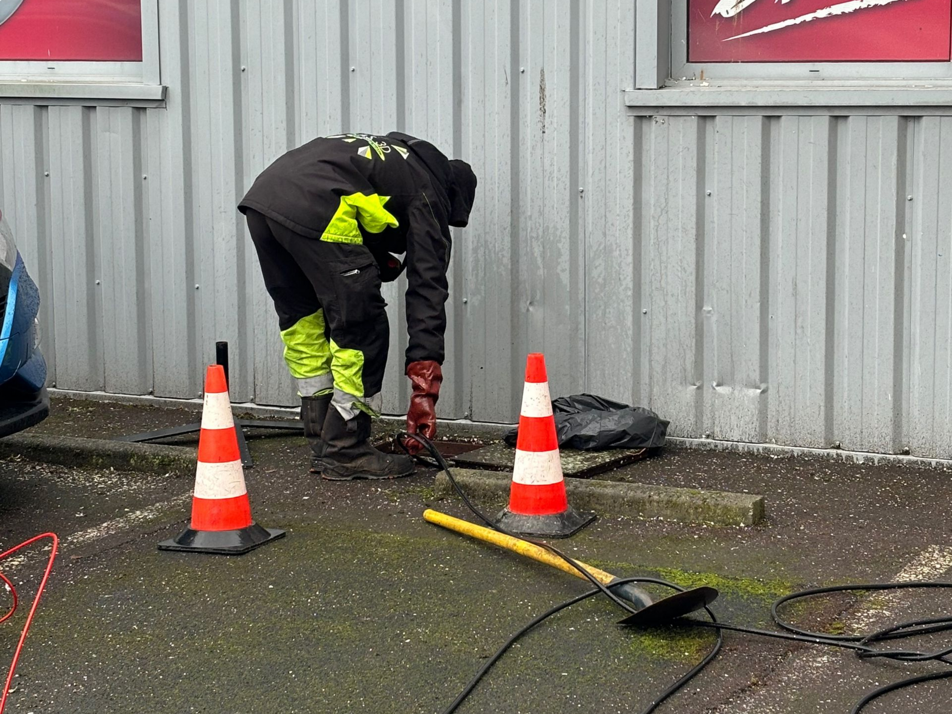 Un ouvrier en tenue de sécurité répare des câbles à l'extérieur d'un bâtiment ; des cônes orange délimitent la zone.