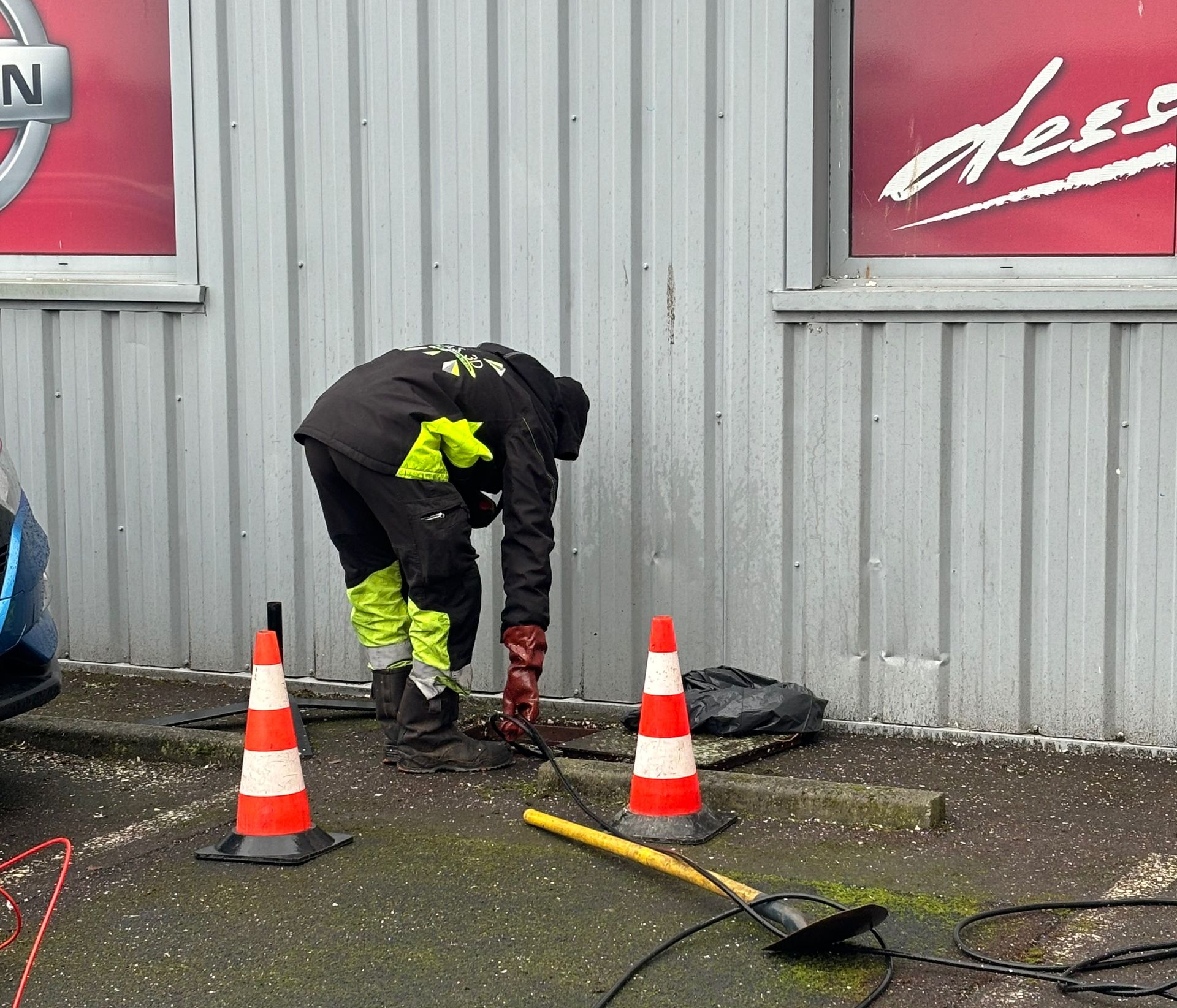 Une personne portant une tenue de travail  à haute visibilité travaille près d'un bâtiment, à proximité de deux cônes de signalisation et d'une pelle.