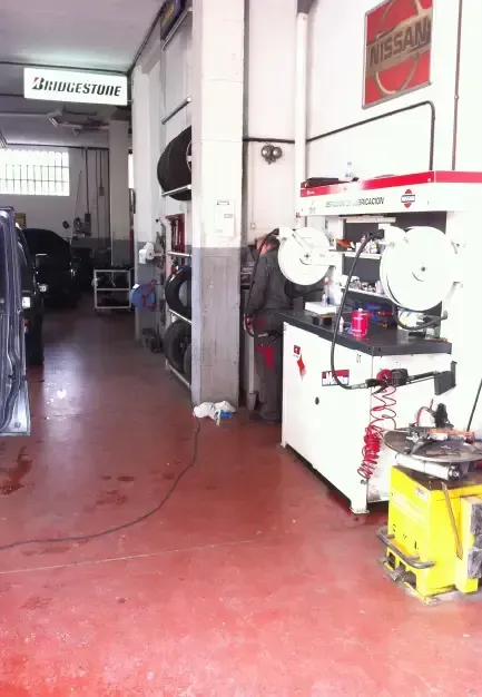 Mecánico trabajando en un garaje; piso rojo, neumáticos, maquinaria, cartel de Nissan, cartel de Bridgestone y coche.