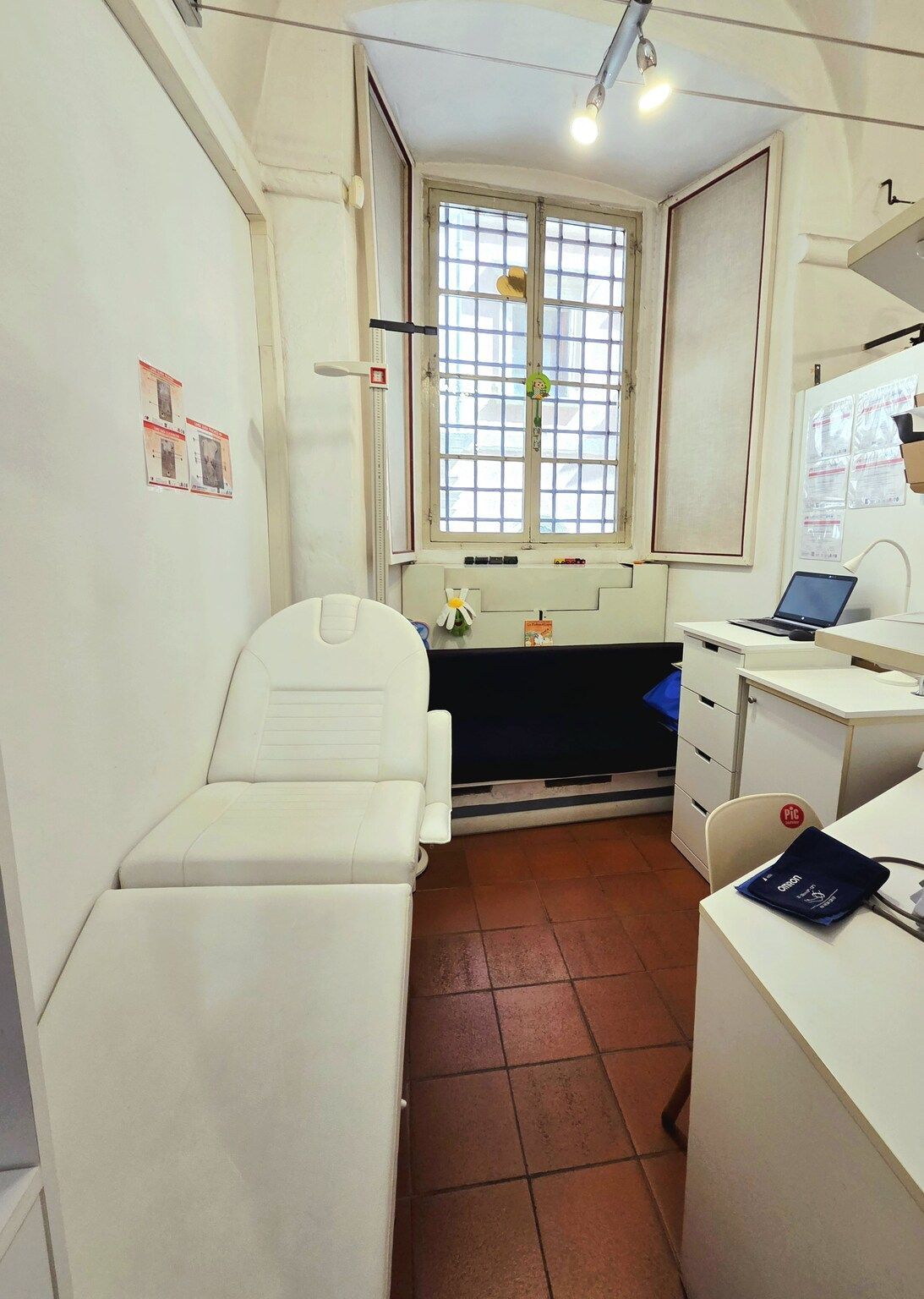Piccola stanza clinica con armadietti bianchi, una finestra con sbarre e un lettino da visita nero.