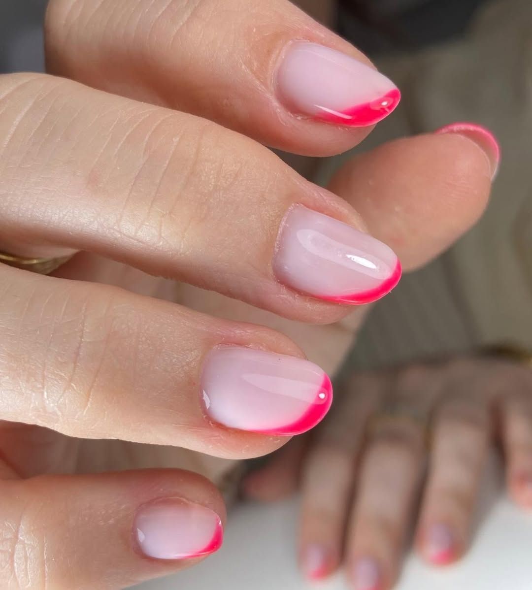Primer plano de uñas bien cuidadas con esmalte rosa pálido y puntas rosa brillante, con una mano borrosa al fondo.