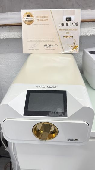 Dispositivo de belleza blanco con detalles dorados y pantalla táctil, expuesto junto a cajas de productos sobre una mesa.