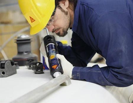 Un homme en salopette bleue, casque jaune et gants, applique du mastic avec un pistolet à calfeutrer sur une structure métallique