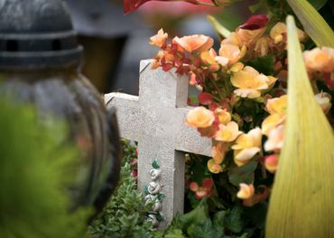 Croix commémorative blanche parmi les fleurs et la verdure, près d'une lanterne sombre dans un cimetière.