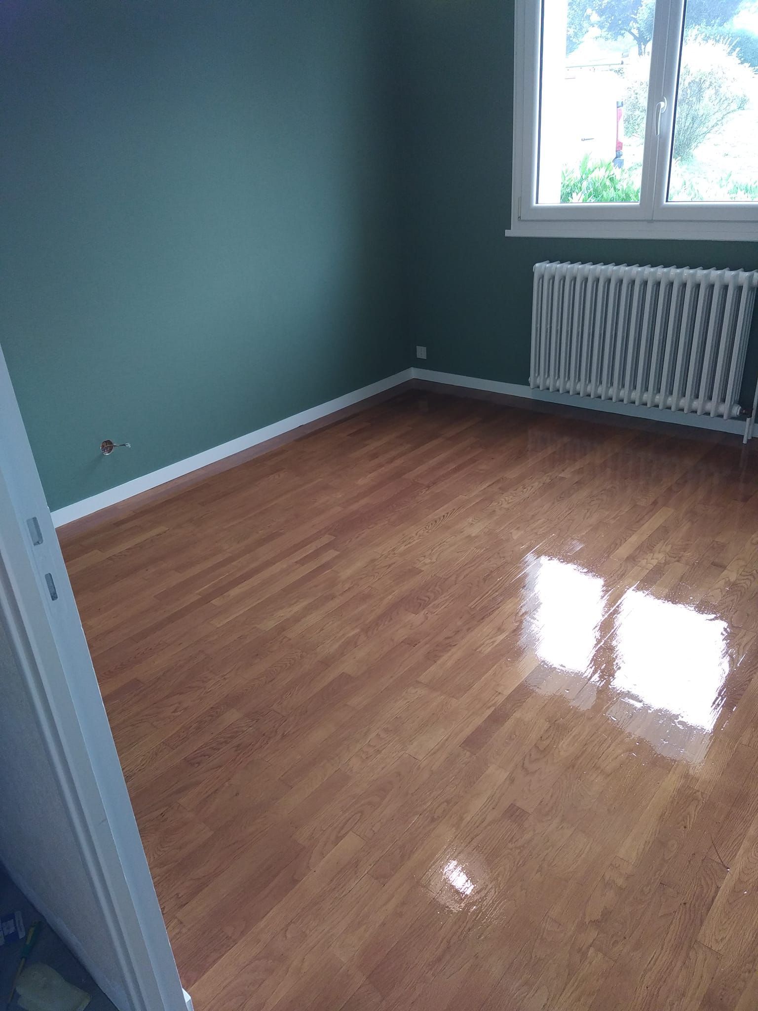 Chambre vide avec plancher de bois, murs verts, fenêtre et radiateur.