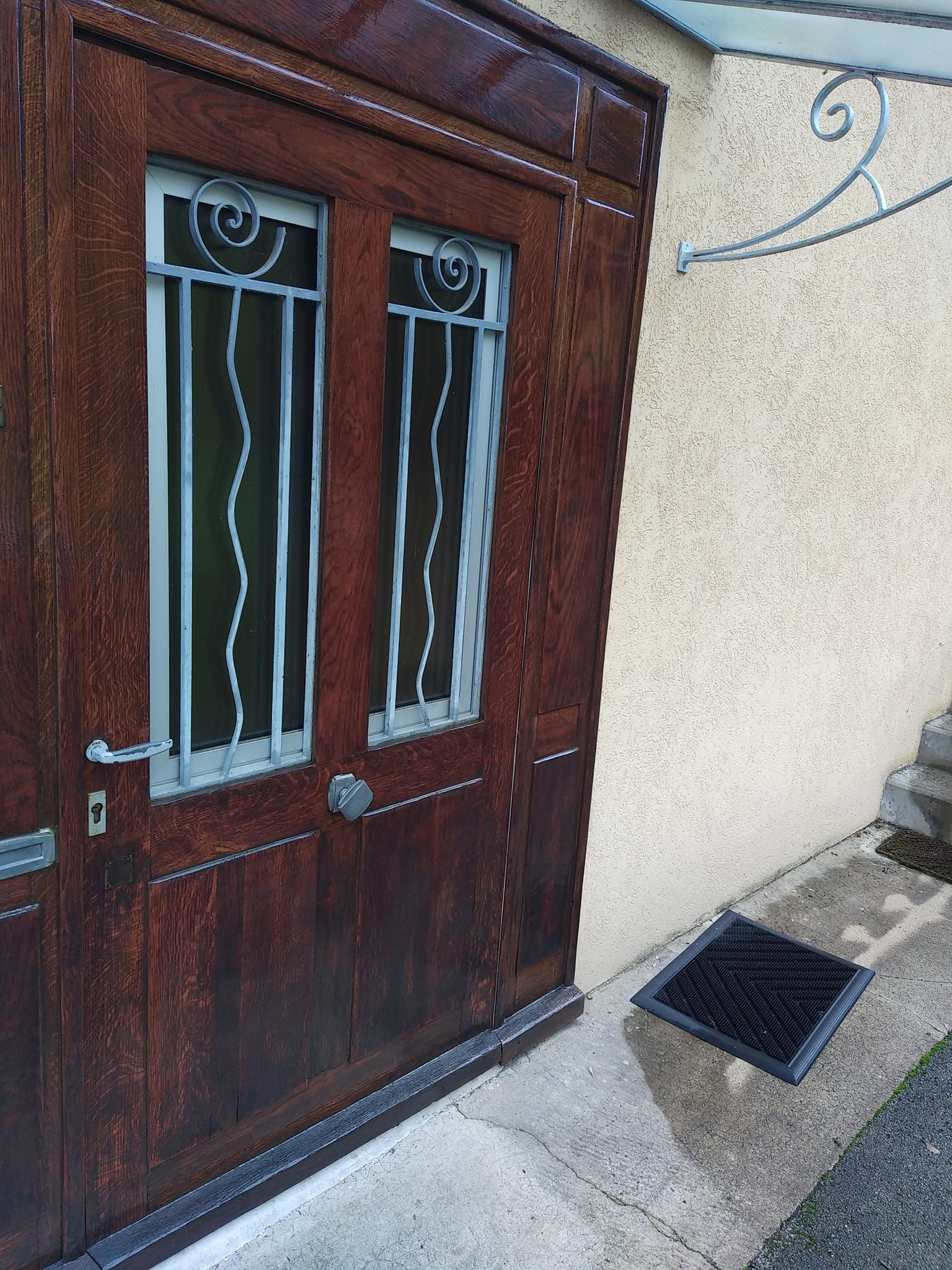 Porte en bois marron avec deux panneaux de verre, ferronnerie ornée et paillasson.