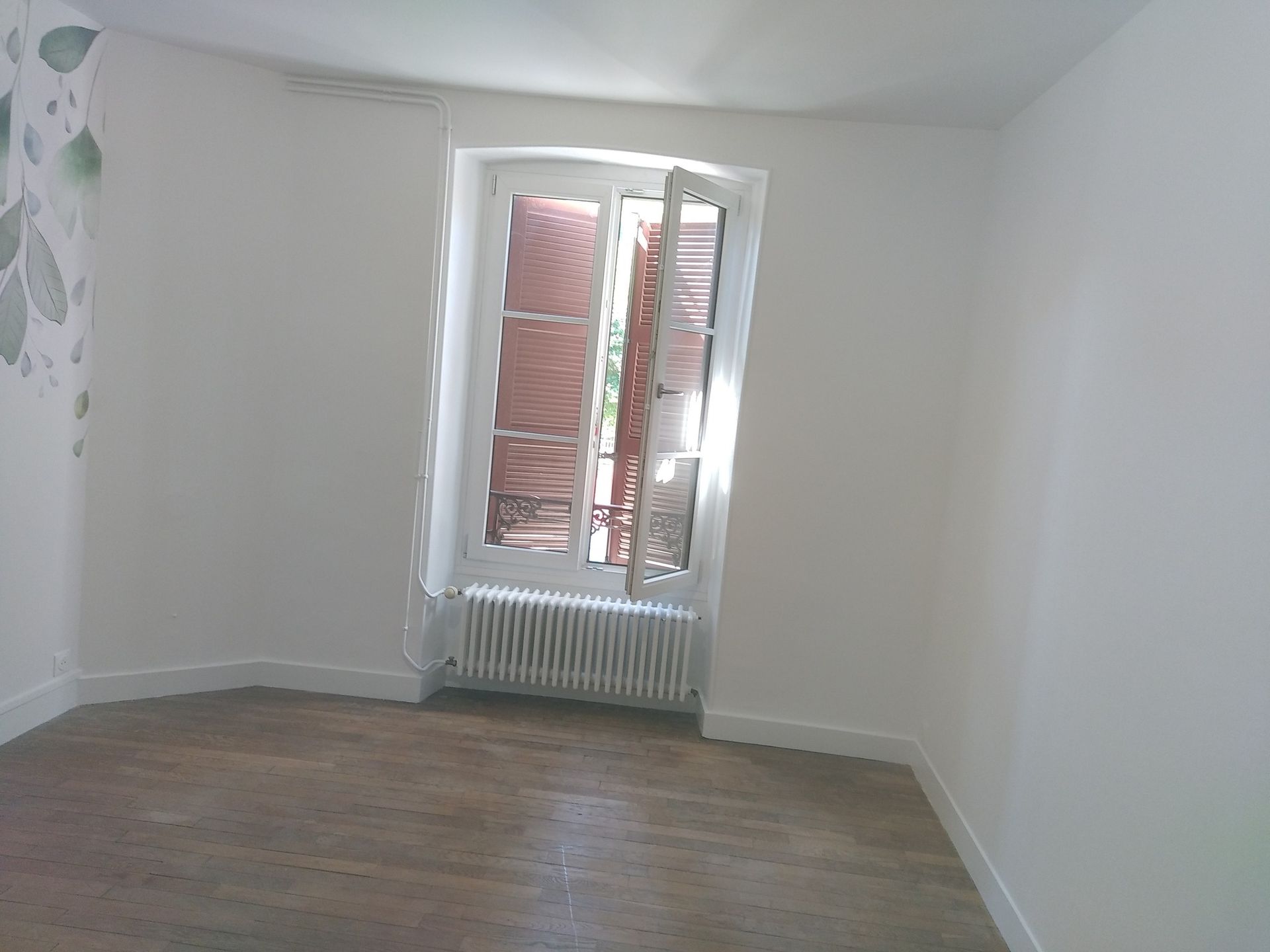 Pièce vide avec parquet, murs blancs et une fenêtre avec rideaux ouverts.