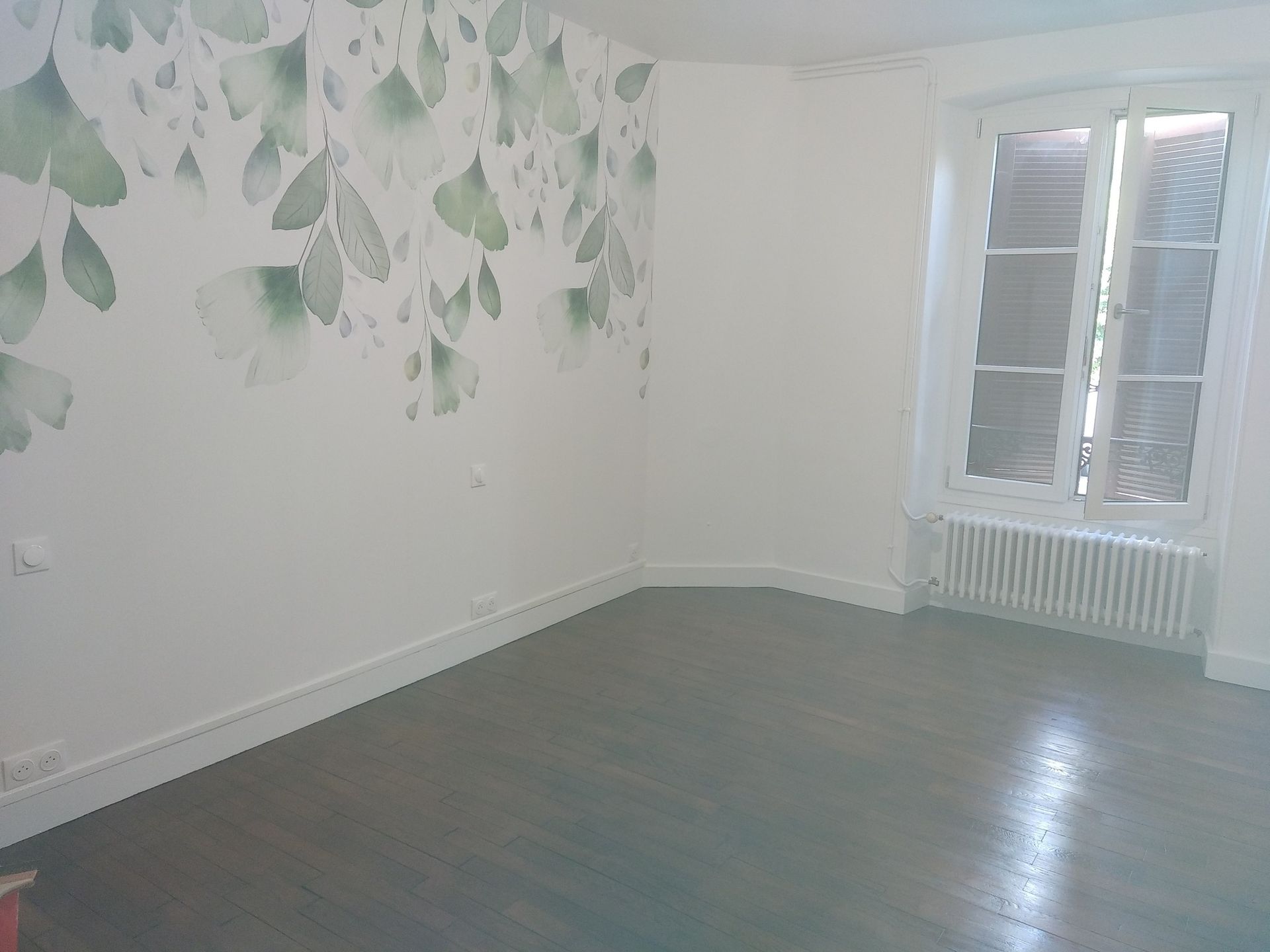 Pièce vide aux murs blancs, au sol gris-bleu et à la grande fenêtre. Un mur est orné d'un motif de feuilles vertes.