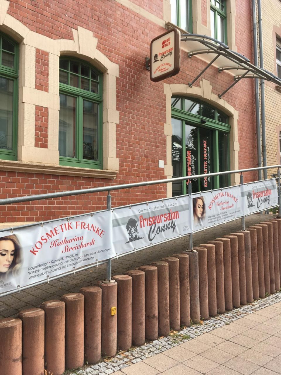 Gebäudeeingang mit Rampe, Beschilderung für ein Kosmetikstudio, rote Backsteinfassade, grüne Zierelemente.