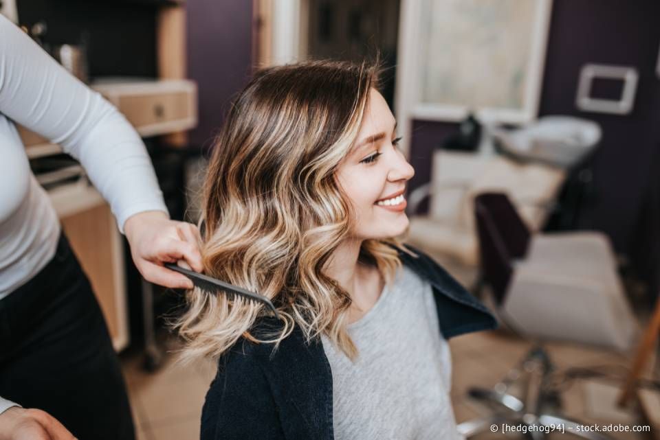 Eine Frau lächelt in einem Salon, während eine Stylistin ihr welliges, gesträhntes Haar kämmt.