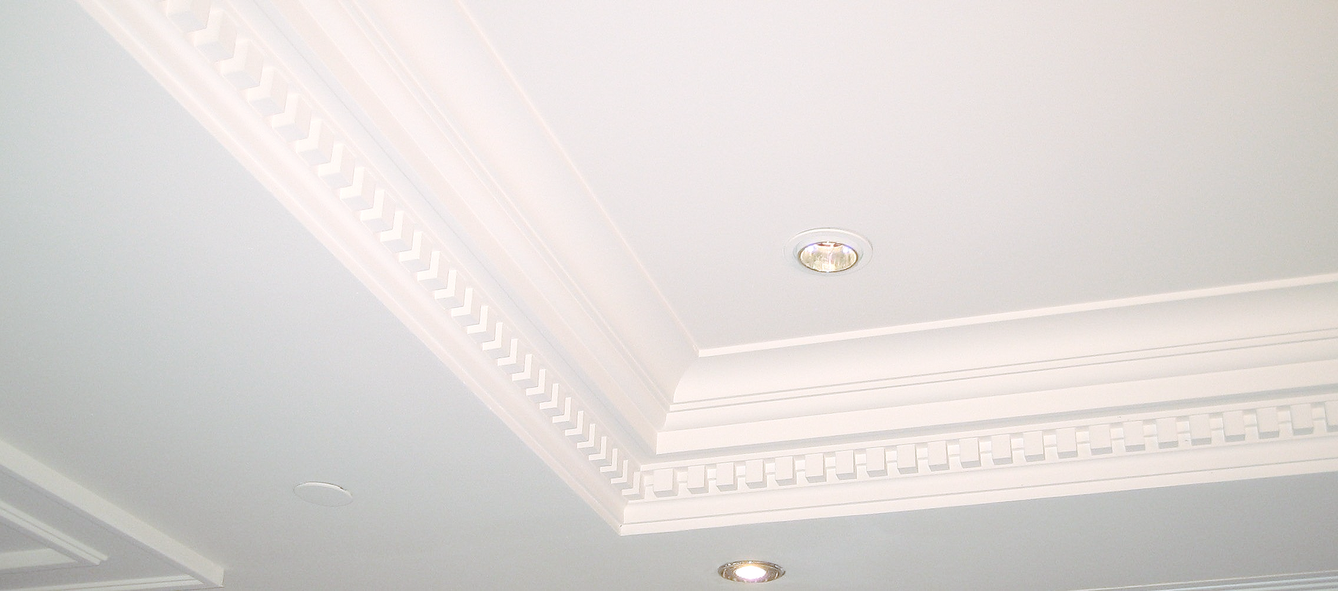 Un coin de plafond blanc orné de moulures décoratives et ouvragées, avec des spots encastrés.