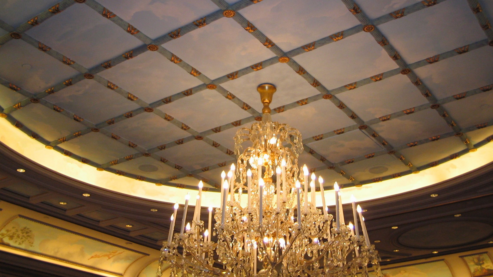 Un grand lustre en cristal orné est suspendu sous un plafond à caissons peint de doux nuages ​​bleus.