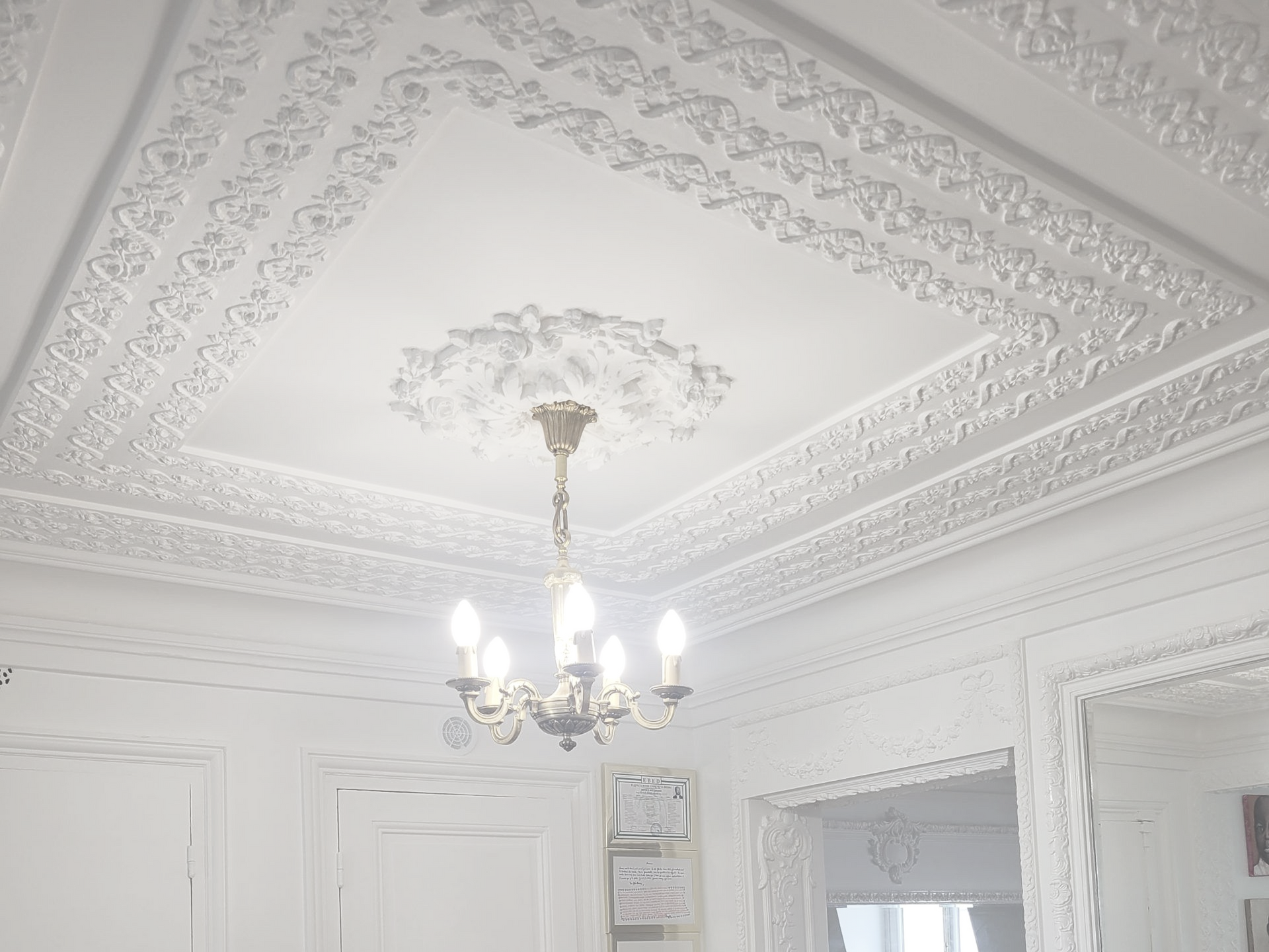 Un plafond blanc orné de moulures complexes et d'un lustre central, vu d'en bas dans une pièce lumineuse et élégante.
