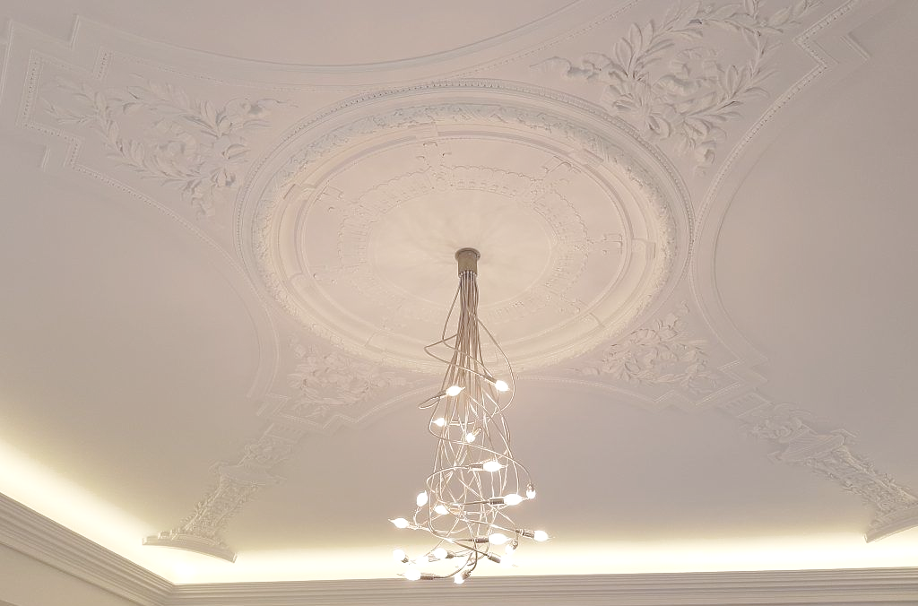 Un plafond blanc décoratif orné de moulures en plâtre est surmonté d'un luminaire moderne en forme de cascade.