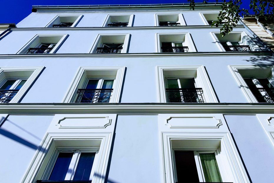 Vue en contre-plongée d'un immeuble bleu clair à plusieurs étages, avec des fenêtres symétriques et des boiseries.