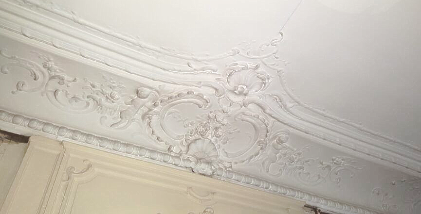 Une moulure de plafond en plâtre blanc finement travaillée, ornée de volutes et de motifs floraux en relief, dans une pièce.