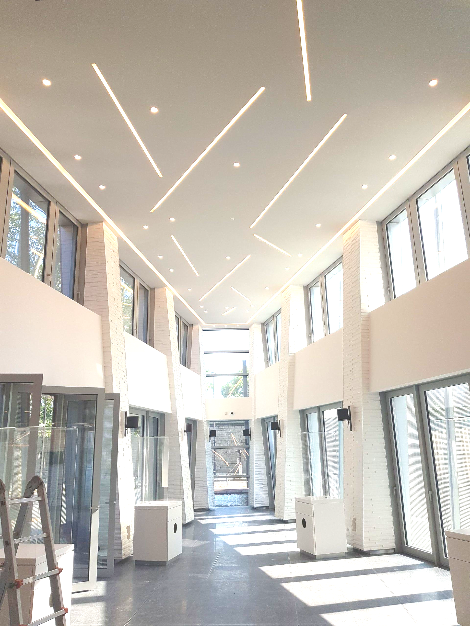 Un couloir moderne et lumineux avec de hautes fenêtres, des colonnes en briques blanches et des bandes lumineuses au plafond.