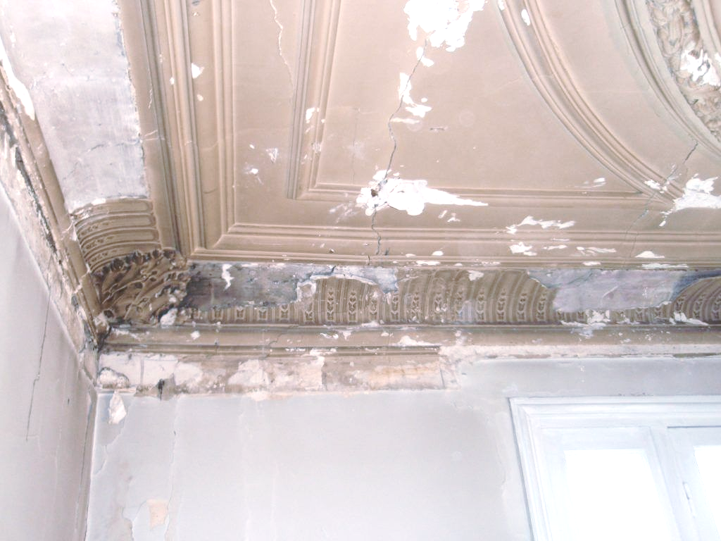Un plafond en plâtre beige orné, abîmé, avec de la peinture qui s'écaille, des fissures.