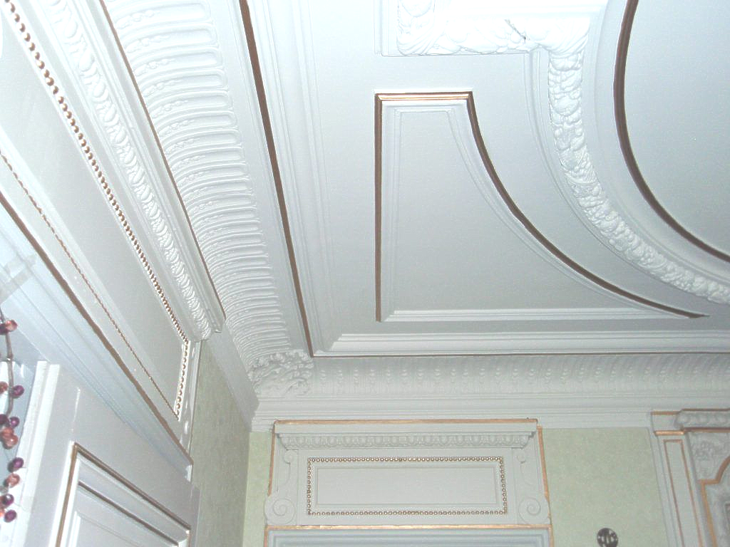 Moulures de plafond blanches ouvragées avec des panneaux encastrés à bordure dorée et des détails floraux complexes.