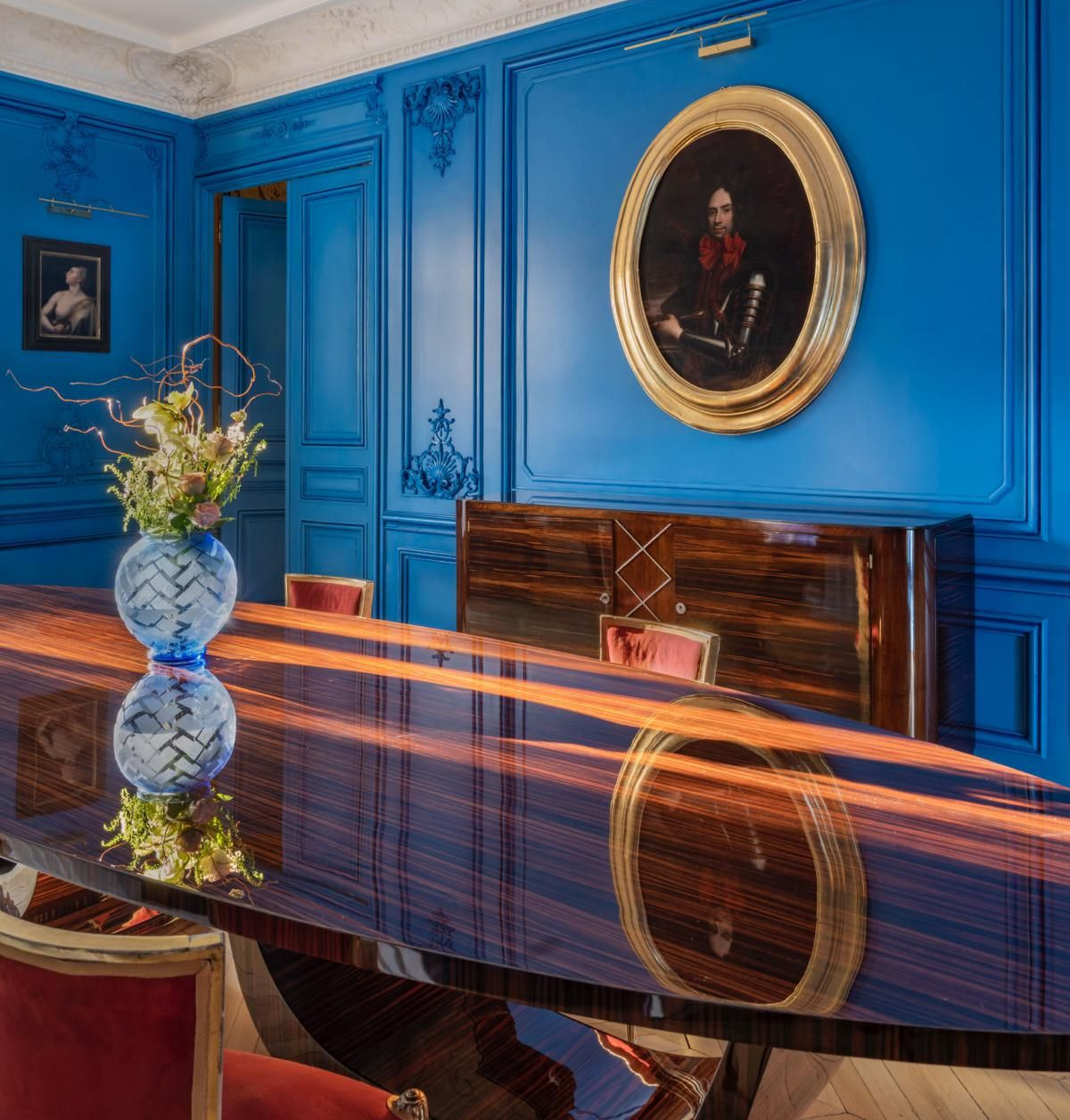 Une salle à manger aux murs lambrissés d'un bleu éclatant, une grande table en bois verni et un portrait dans un cadre ovale.