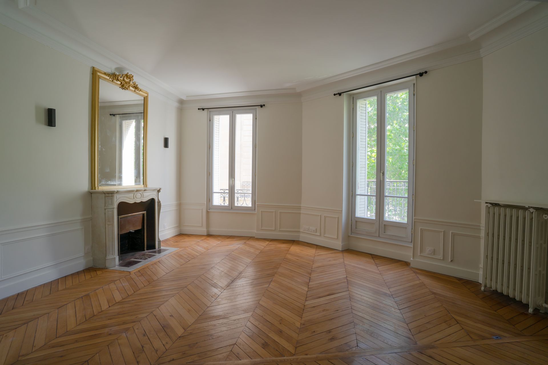 Une pièce vide , au parquet en chevrons, avec une cheminée surmontée d'un miroir doré et de deux hautes fenêtres.