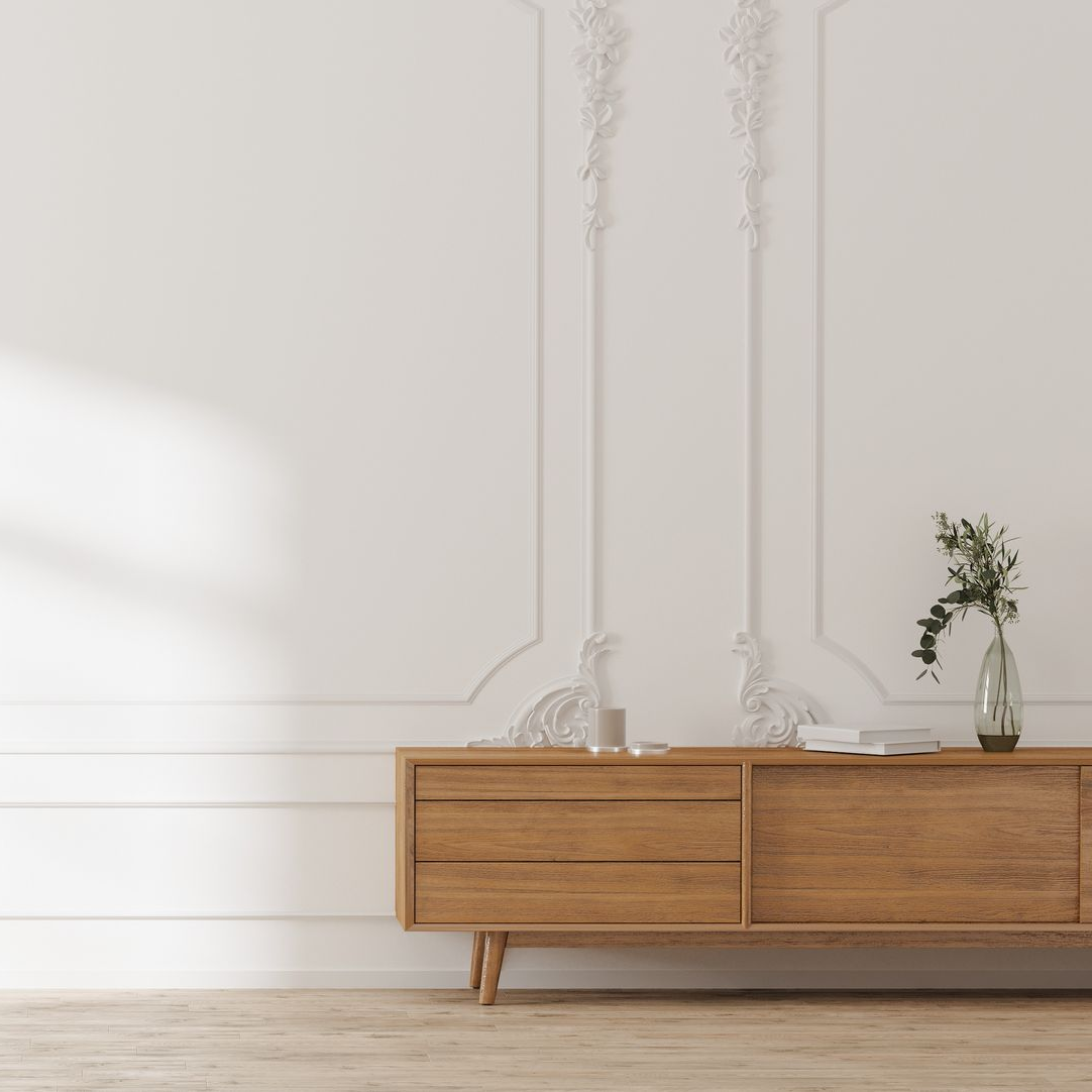 Un meuble en bois, surmonté d'un vase en verre et de livres, est adossé à un mur blanc orné de moulures classiques.